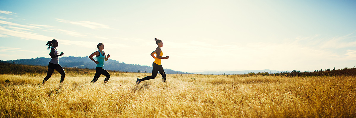 A group of young adult friends runs in a pristine and vast outdoor area, the sun shining warm golden light on the scene. They run across a large field, the grass a golden yellow color. Shot in a minimalist style. Depicting trail running, healthy lifestyle and exercise in the great outdoors.