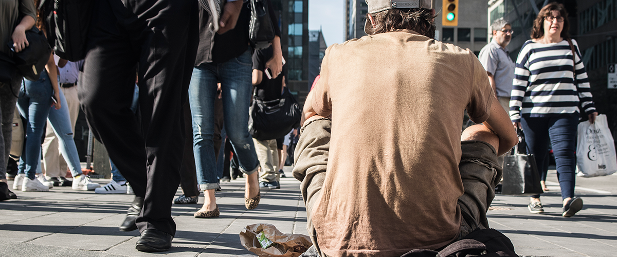 Un sans-abri avec un sac à dos assis sur le trottoir d’une rue achalandée du centre-ville de Toronto, entouré de passantes et passants